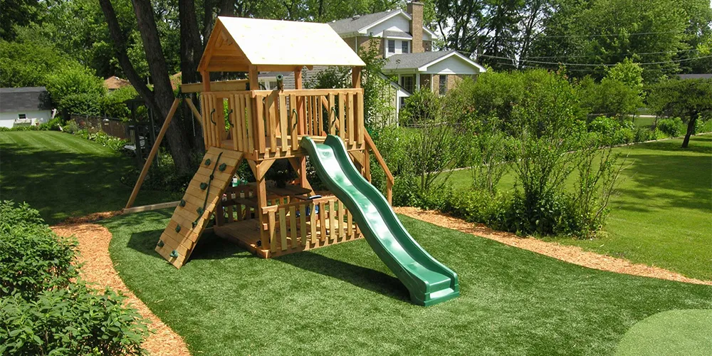 A children's play area with a wooden playset on synthetic grass, surrounded by natural landscaping, providing a safe and clean space for outdoor fun.